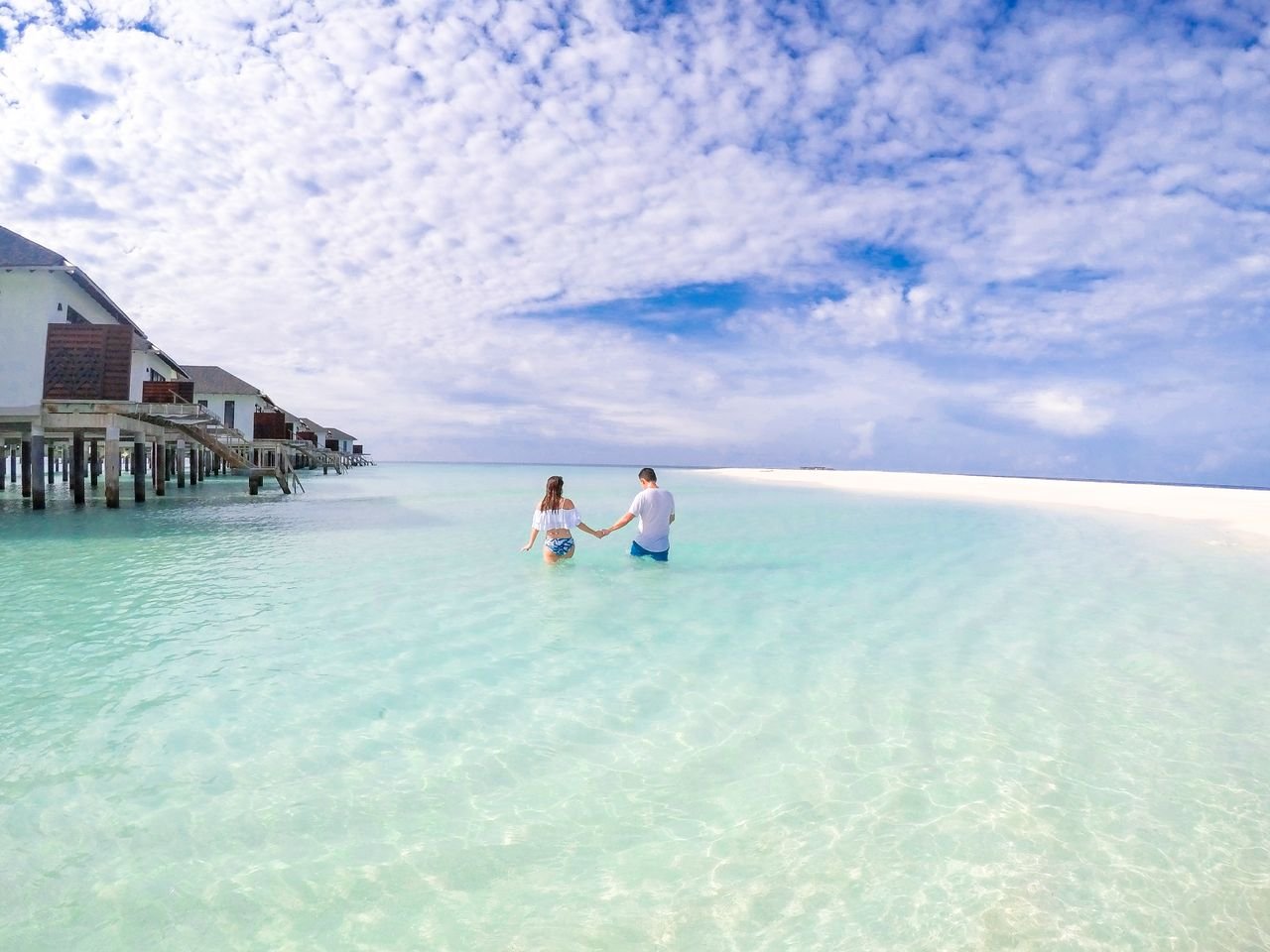 Home A couple holding hands while walking in crystal clear ocean waters under a vibrant sky.