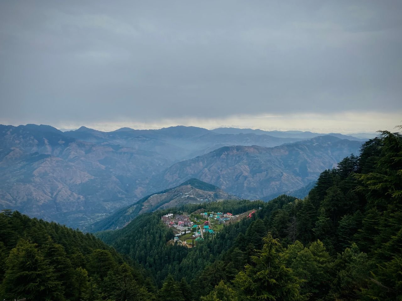 Home A serene view of a village nestled in the evergreen Shimla hills under overcast skies.
