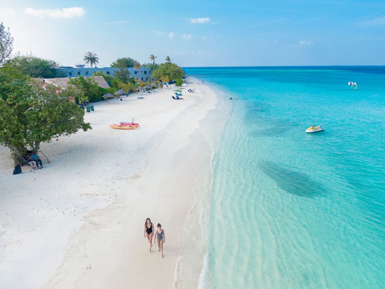 Home Aerial view of two women walking on a beautiful white sand beach in the Maldives with turquoise water.